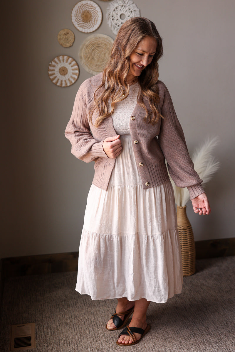Woman wearing a pink cardigan over a white dress in a room with decorative wall art.