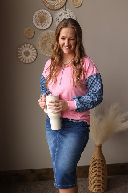 Woman in a pink and blue sweatshirt and midi denim skirt holding a white cup, standing in a room with decorative wall art.