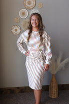 A woman wearing a cream and tan horizontally striped long-sleeve midi dress with a tie waist, standing in a room with a gray wall decorated with circular woven wall art.