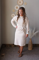 A woman modeling a cream and tan striped long-sleeve knit dress with a tie waist, standing in a styled room with neutral walls, woven wall decor, and a vase with pampas grass.