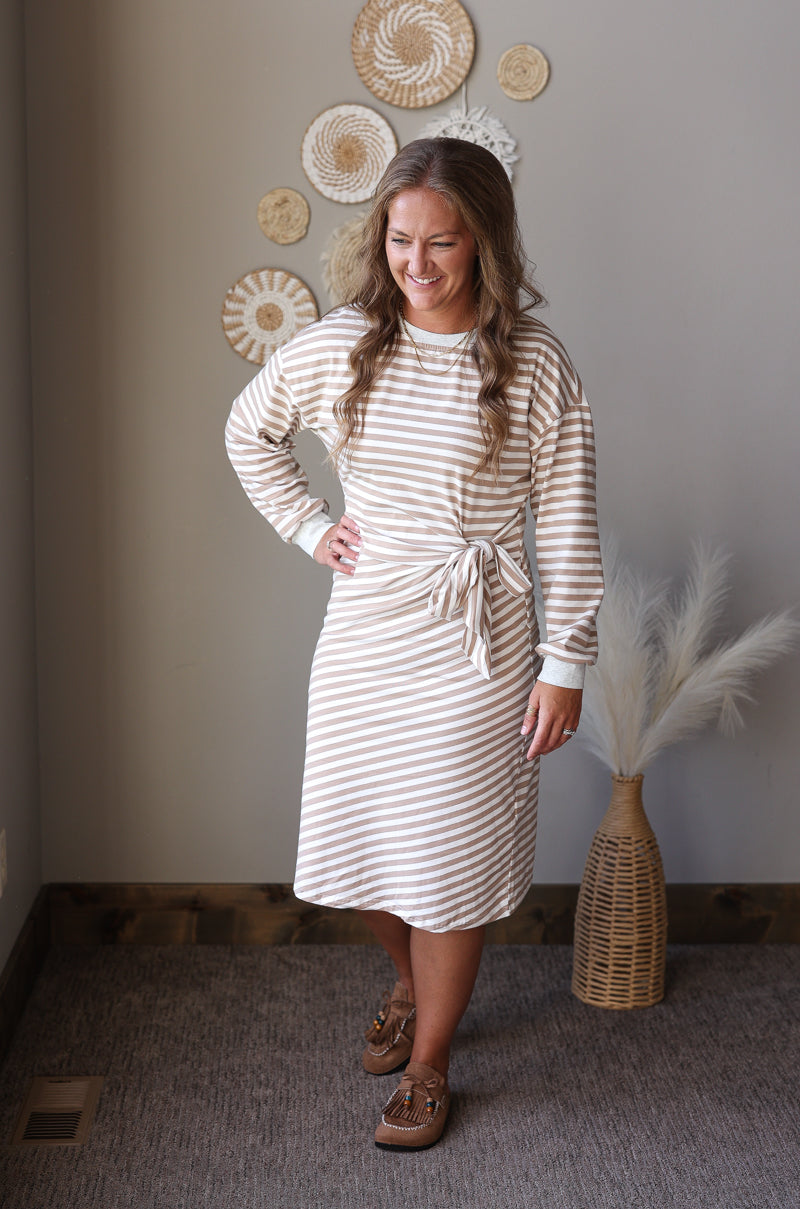 A woman modeling a cream and tan striped long-sleeve knit dress with a tie waist, standing in a styled room with neutral walls, woven wall decor, and a vase with pampas grass.