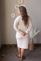A woman wearing a beige and white striped long-sleeve knit dress, shown from the back in an indoor setting with neutral-toned walls and home decor.