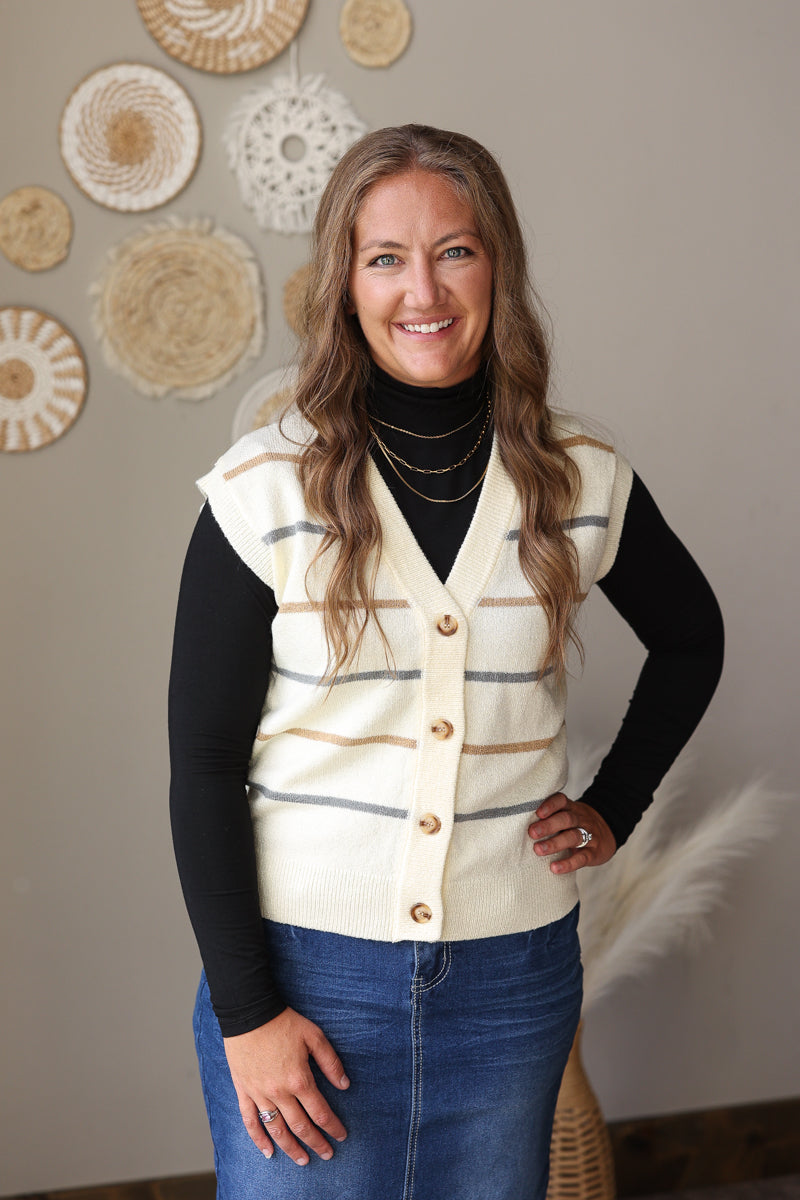 A woman wearing a cream knit vest with horizontal gray and tan stripes over a black turtleneck and blue jean skirt, standing indoors against a beige wall with woven wall decor.