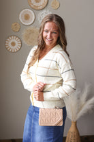 A woman wearing a cream and gray striped knit cardigan sweater and a blue denim skirt, holding a beige quilted crossbody bag. She is standing in front of a neutral gray wall decorated with circular woven wall art.