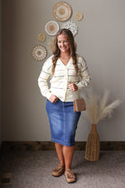 A woman modeling a cream and tan striped knit cardigan, blue denim midi skirt, and brown loafers, standing in a styled room with decorative wall art and a vase of pampas grass.