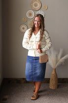 A woman stands indoors wearing a cream striped button-front cardigan, a blue denim midi skirt, tan loafers, and carrying a small quilted tan handbag. The background shows a neutral wall with decorative woven wall art and a wicker vase with pampas grass.