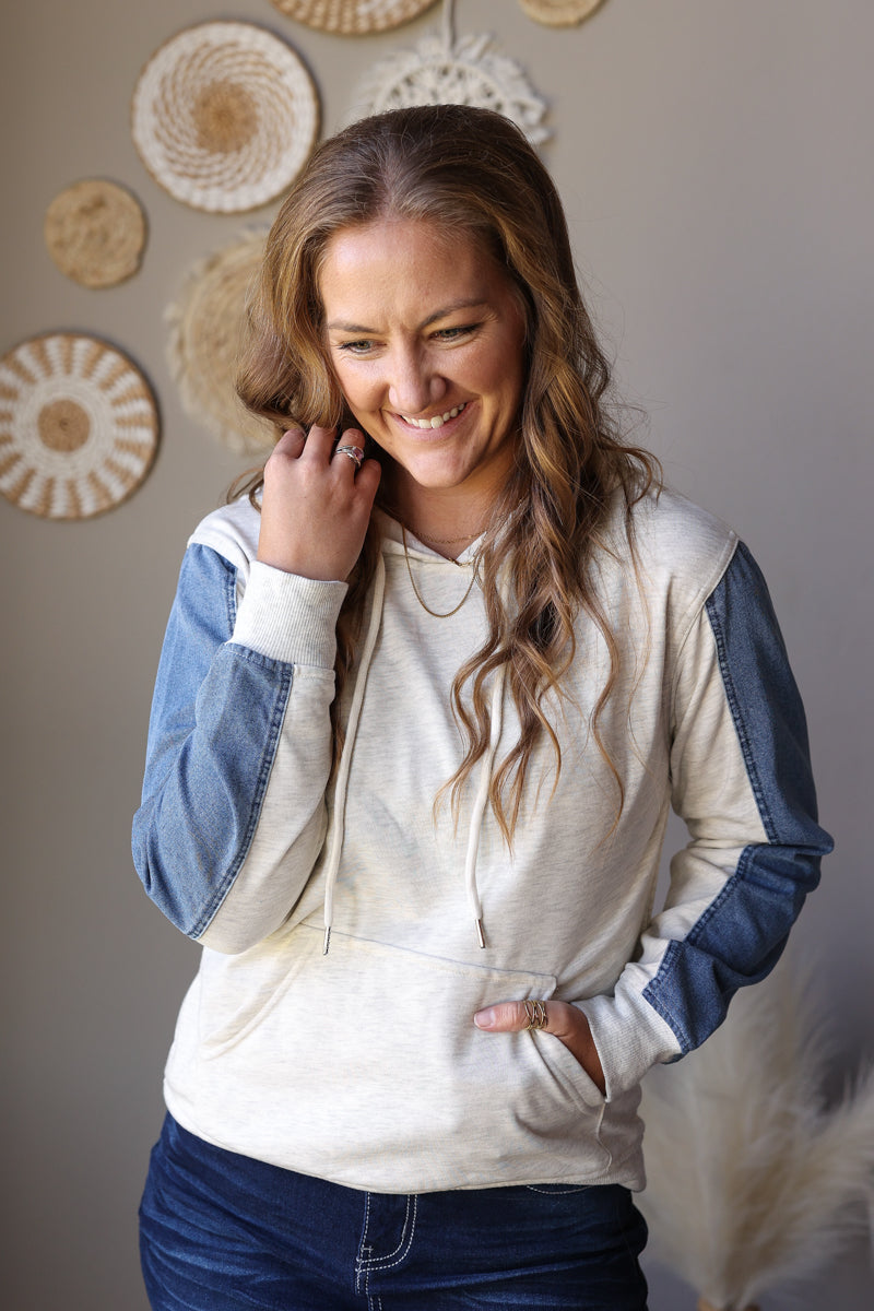 Close-up of a light grey hoodie with blue denim sleeve panels, drawstrings, and a front pouch pocket.
