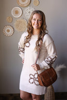 Angled front view of a parchment sweater dress with brown floral outlines and a brown crossbody bag.