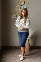 A woman modeling a striped long-sleeve knit sweater and a blue denim midi skirt, standing in a styled room with a neutral wall, decorative woven wall art, and a wicker vase with dried grass.