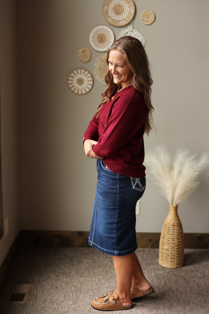 A woman stands in profile wearing a burgundy long-sleeve top and a blue denim skirt, with tan slide sandals, in an indoor room setting with decorative wall art and a vase of pampas grass.