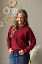 A woman with long wavy light brown hair smiling, wearing a burgundy zip-up knit sweater and a blue denim skirt. She is standing against a neutral beige wall decorated with circular woven wall art.