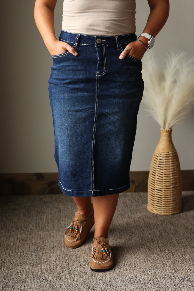 A person wearing a dark blue denim midi skirt with a straight, slightly flared silhouette, paired with a beige top and brown moccasin-style shoes. The setting is indoors on a textured gray carpet with a woven vase of dried grass in the background.