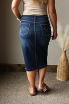 A person shown from the back wearing a dark blue denim pencil skirt with zigzag pocket details, paired with a beige top and brown sandals, standing on a carpeted floor next to a woven vase with dried grass.