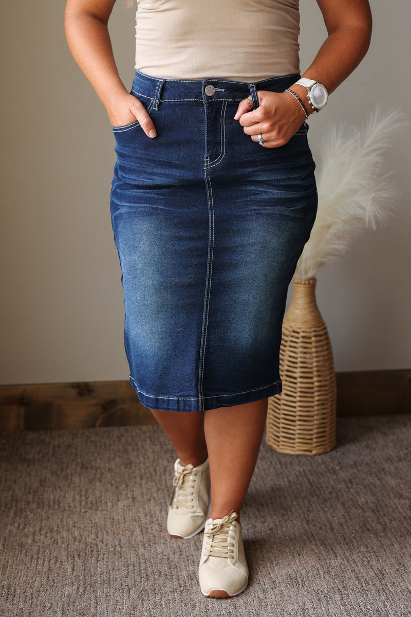 A person wearing a dark blue denim pencil skirt with rolled hems, paired with a beige top and beige sneakers, standing in an indoor setting with a woven vase in the background.