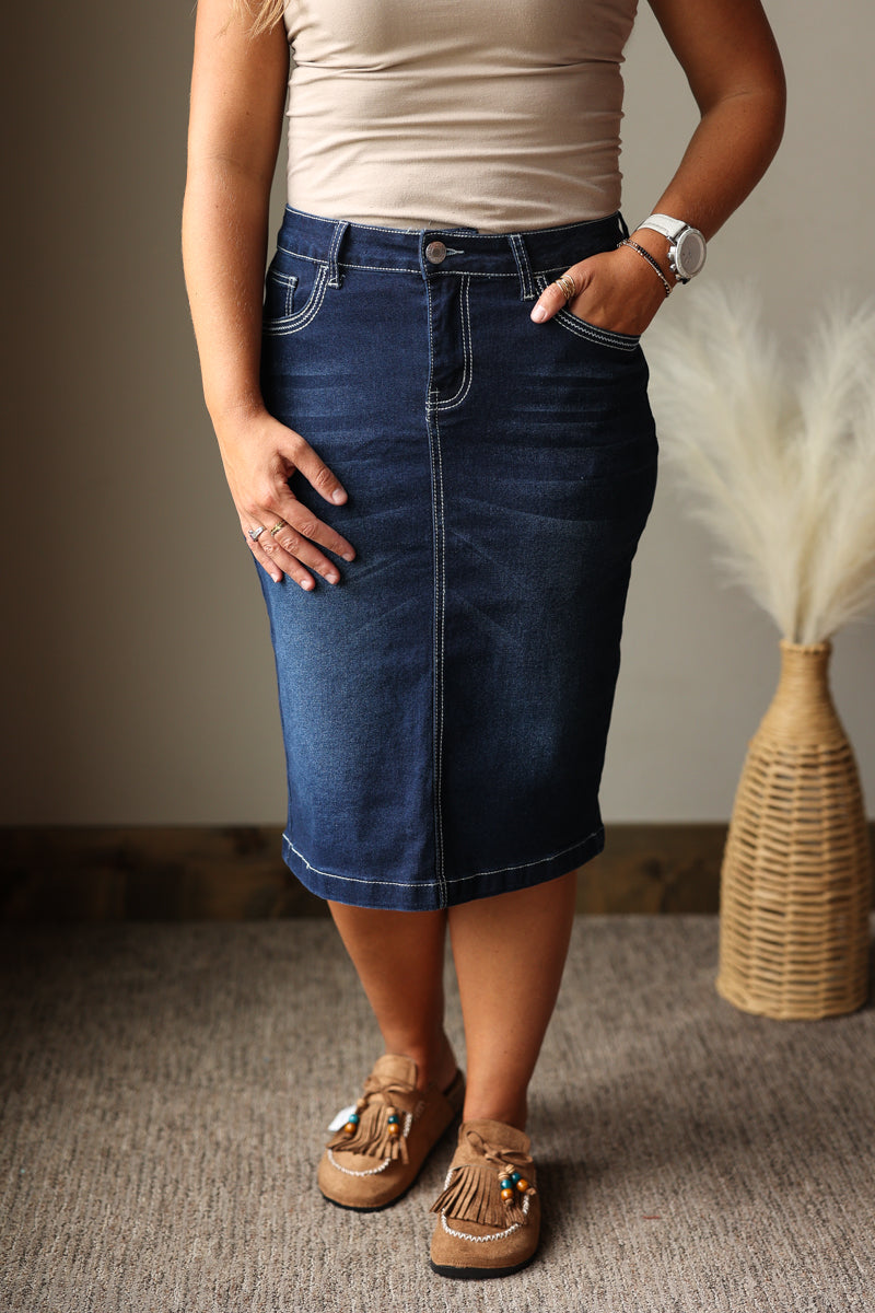 A person wearing a dark blue denim pencil skirt with contrast white stitching, paired with a beige top and brown mule sandals, standing in an indoor setting.