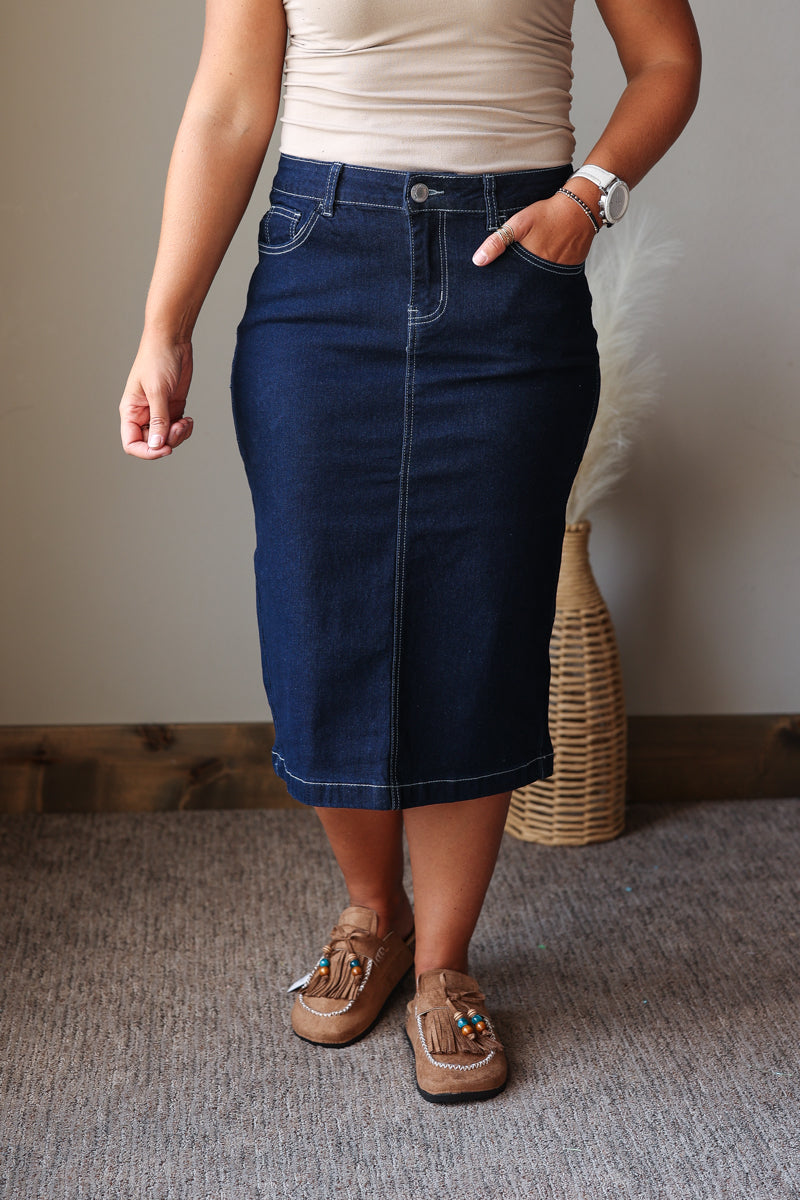 A person wearing a dark blue denim midi skirt, paired with a beige top and tan moccasin-style shoes, standing in an indoor setting with a wicker basket and neutral wall in the background.