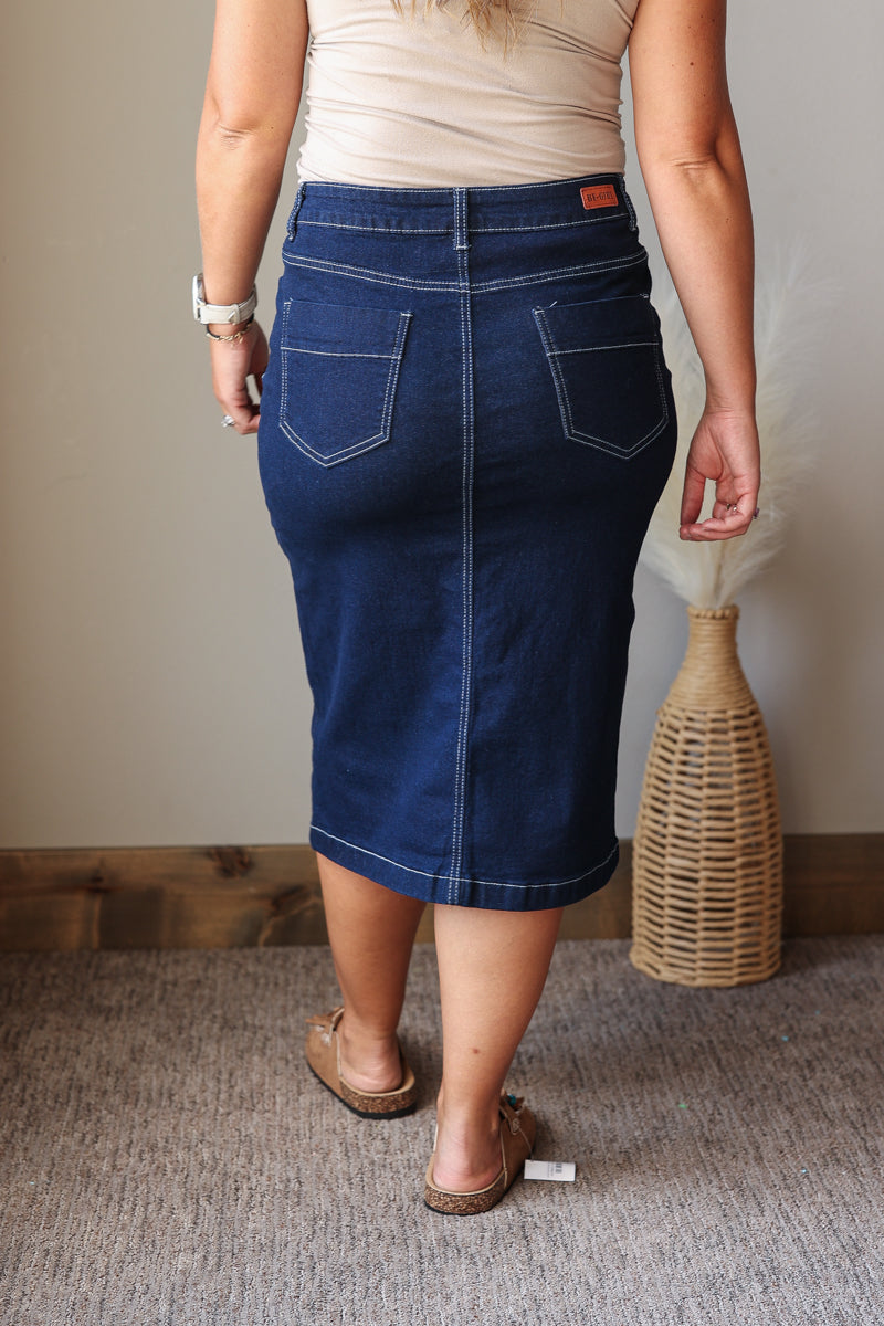 A woman shown from the back wearing a dark blue denim pencil skirt with white contrast stitching and two back pockets, paired with a beige top and brown sandals in an indoor setting.