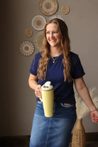 Woman holding a yellow mug in a room with decorative wall art wearing a navy crew essential tee shirt