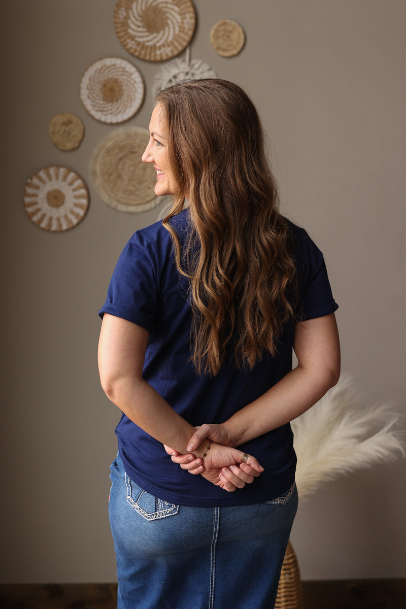 Woman wearing a blue shirt and jean skirt standing in front of decorative wall art.
