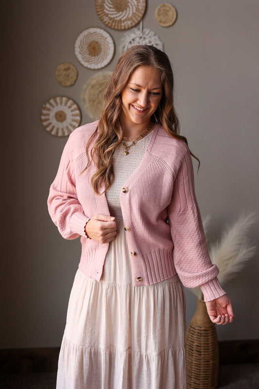 Woman wearing a pink cardigan over a white dress in a room with decorative plates on the wall.
