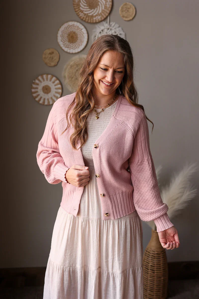 Woman wearing a pink cardigan over a white dress in a room with decorative plates on the wall.