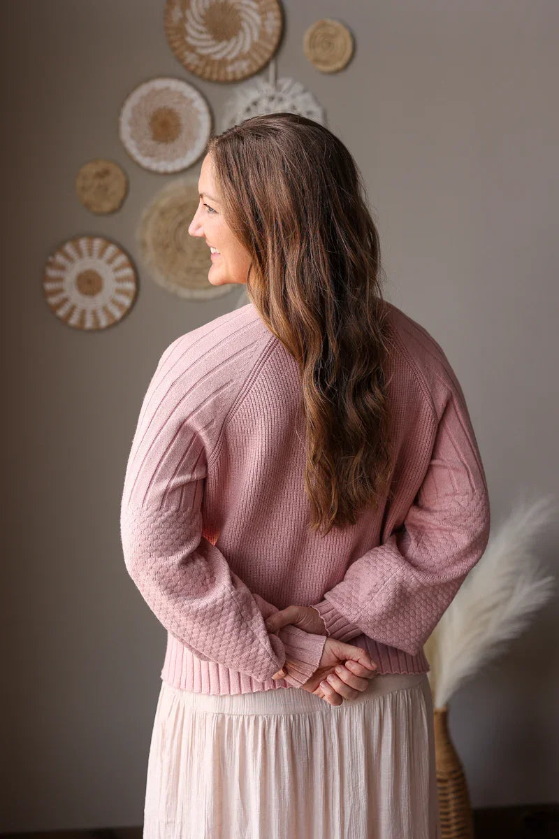 Woman wearing a pink sweater and white skirt standing in a room with decorative wall art.