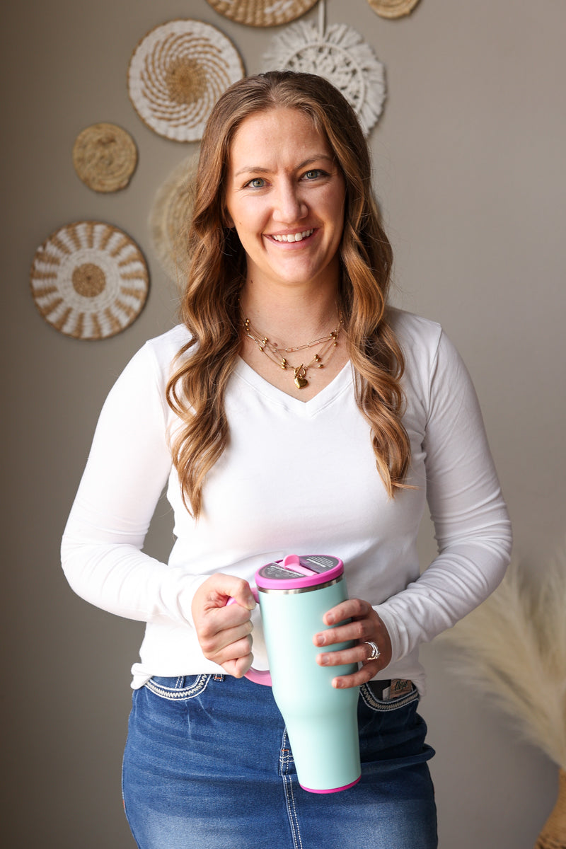 Woman holding a light blue mug with a pink rim in a home setting, wearing a white vneck long sleeve layer top.
