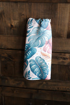 A hanging geometry-like tea towel with a tropical leaf pattern in teal, blue, pink, and beige, displayed against a dark wood plank background.