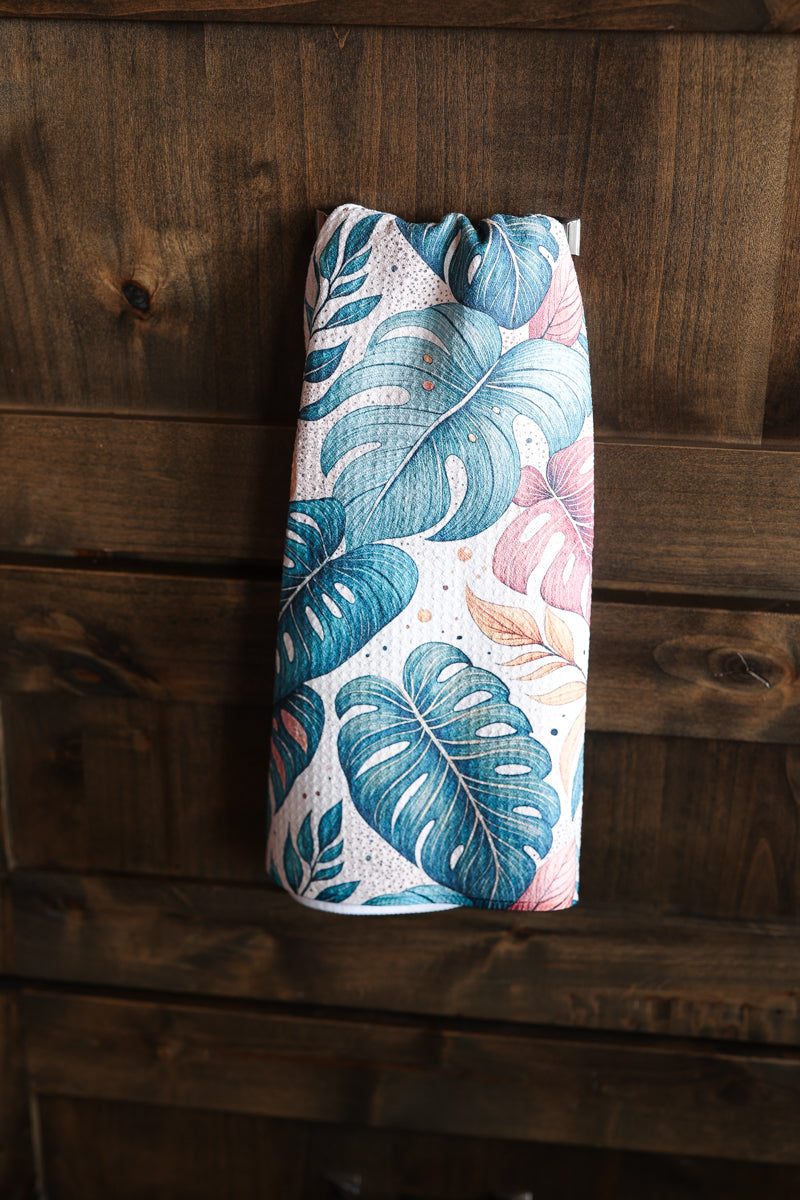 A hanging geometry-like tea towel with a tropical leaf pattern in teal, blue, pink, and beige, displayed against a dark wood plank background.