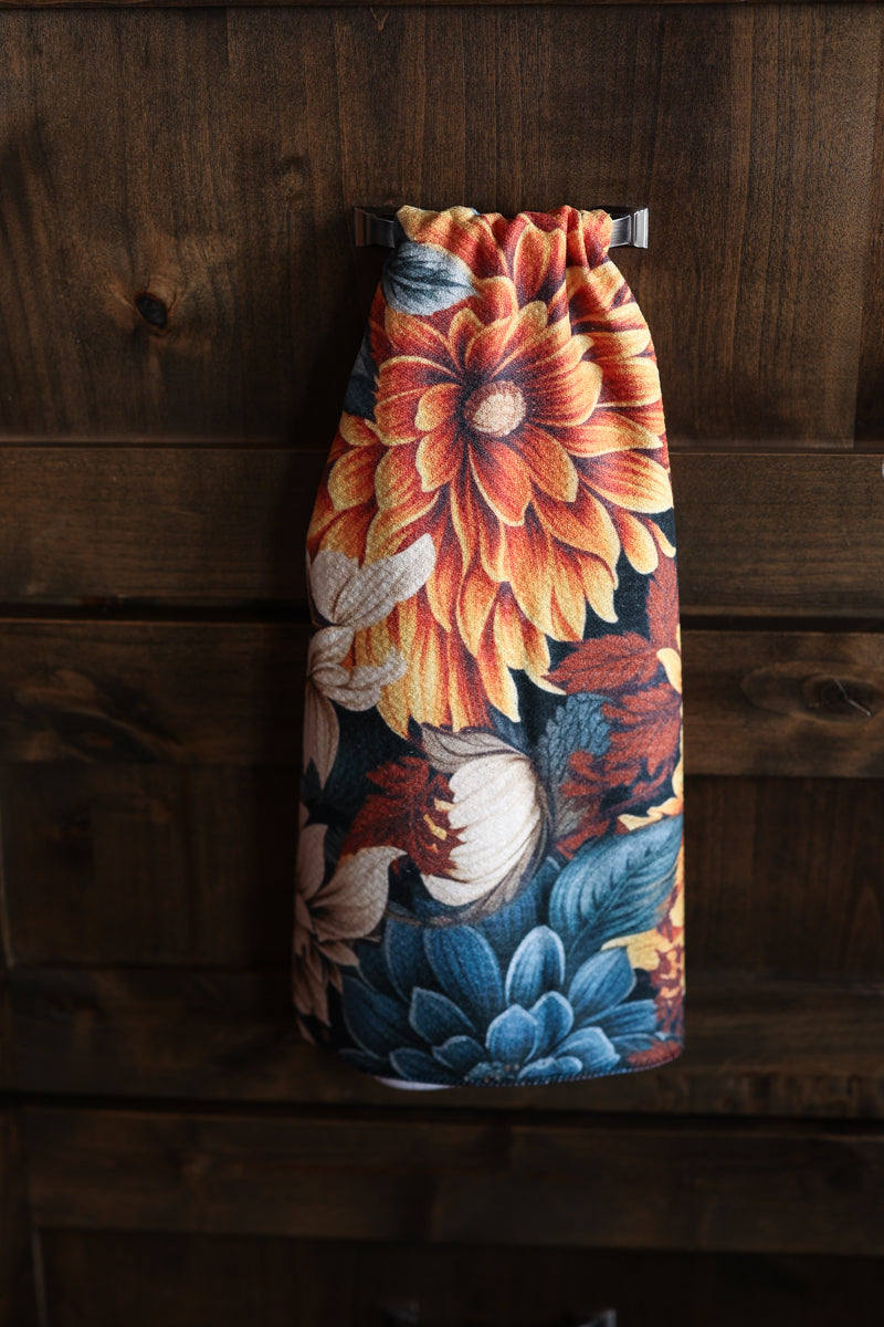 A drawstring bag with a vibrant floral print featuring a large orange dahlia, blue flowers, and white blossoms on a dark background, hanging against a dark wood plank wall.