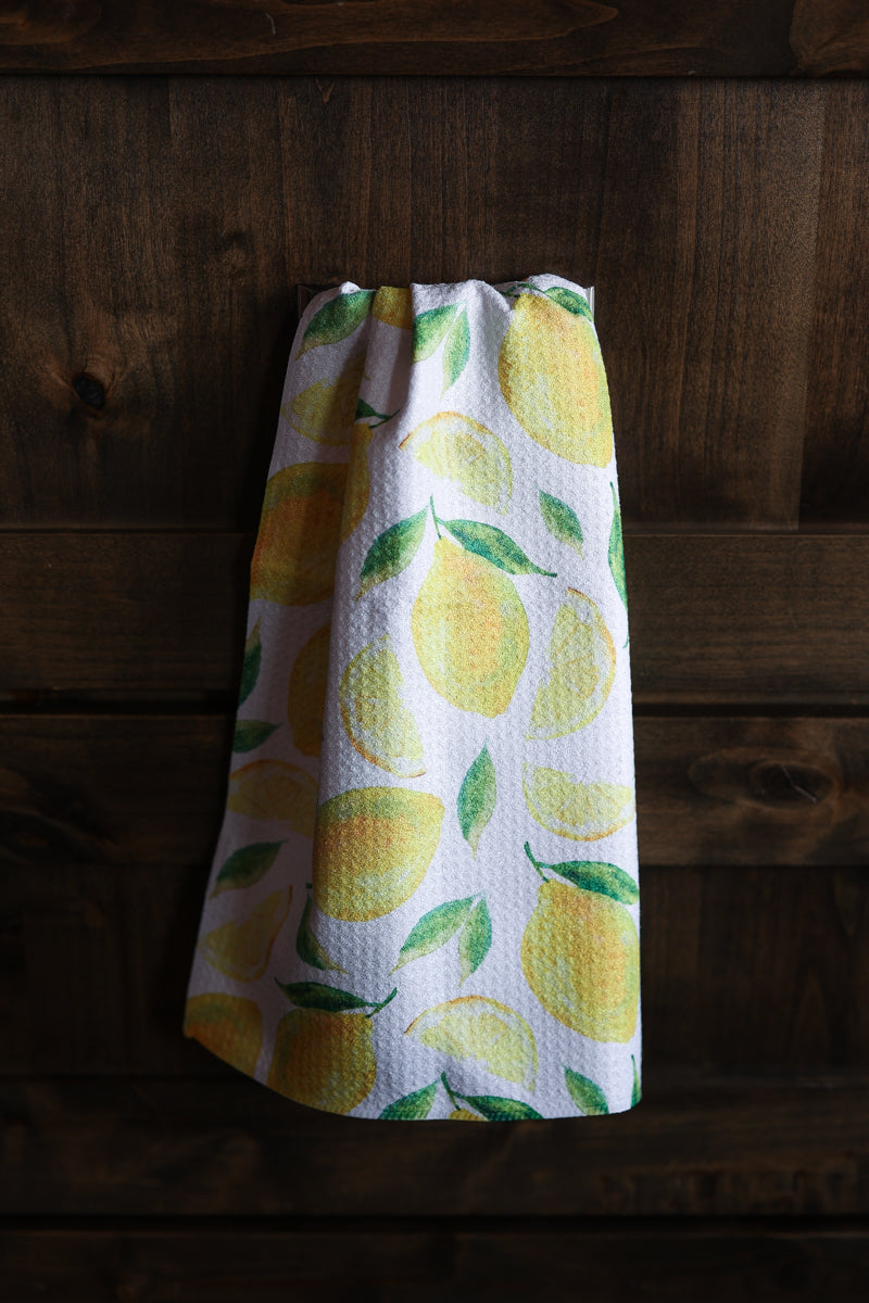 A white kitchen towel with a yellow lemon and green leaf pattern, hanging against a dark rustic wooden plank background.