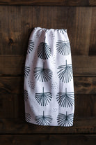A white kitchen towel with a black palm frond pattern, hanging against a dark wood plank background.