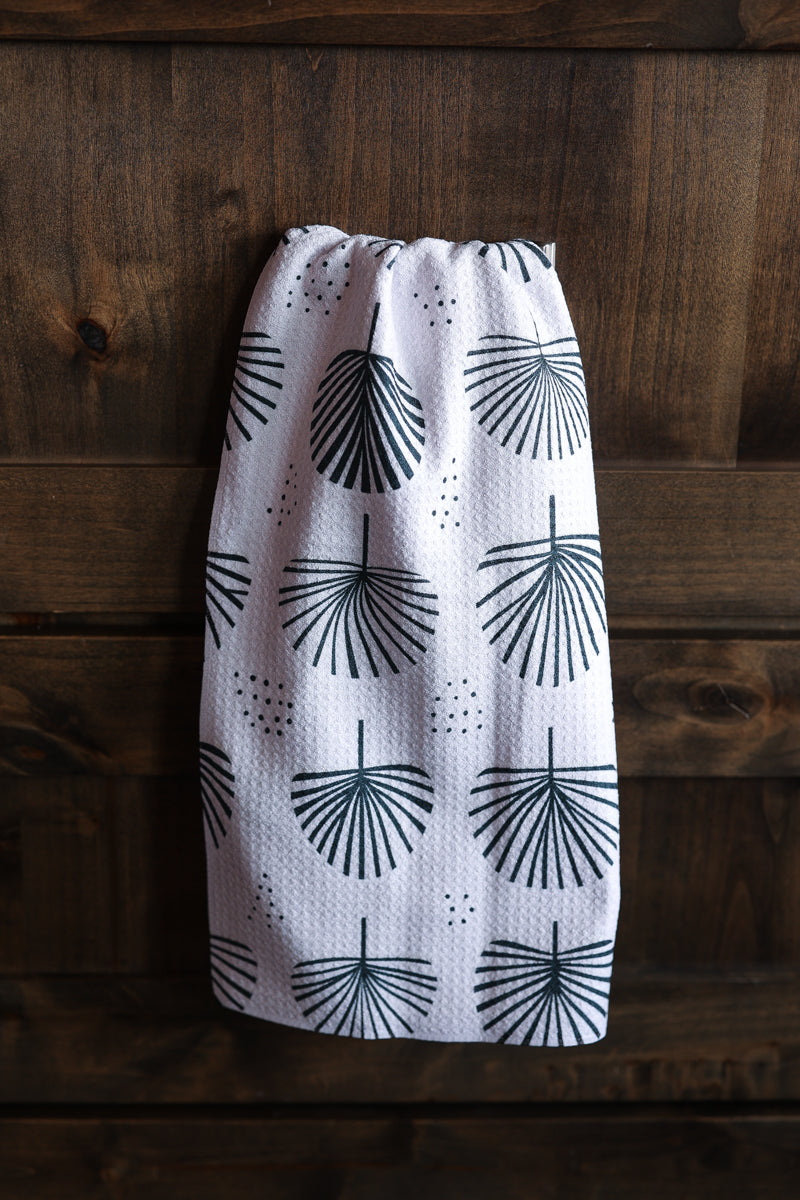 A white kitchen towel with a black palm frond pattern, hanging against a dark wood plank background.