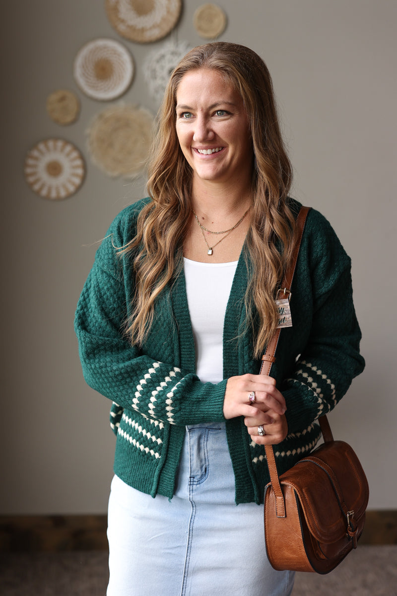 Emerald green textured cardigan with cream diamond stripe cuffs and hem, worn open over a white top and light denim skirt.