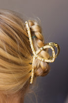 A close-up of a blonde braid secured with a gold claw hair clip decorated with small clear rhinestones, shown against a dark gray background.