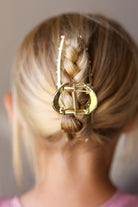 A close-up back view of a person wearing a large gold claw-style hair clip holding a braided low ponytail.