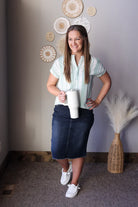 Woman holding a white mug in a room with decorative wall art and a vase wearing a green white striped blouse and black denim midi skirt