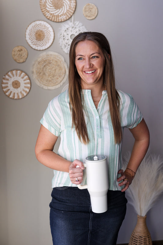 Woman holding a white mug with decorative wall art in the background wearing a green white striped vneck henley blouse