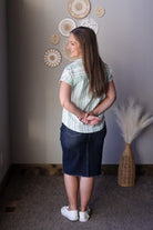 Woman standing in a room with decorative wall items and a vase wearing a green white striped blouse and black denim skirt