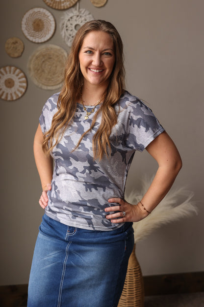 Woman wearing a camouflage t-shirt and blue jean skirt standing in a room with decorative wall items.