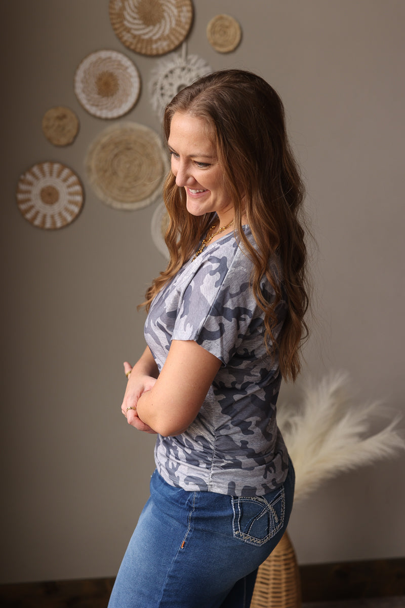 Woman standing with arms crossed in a room with decorative plates on the wall, wearing a grey camo summer top and denim skirt