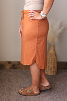 A person wearing a heathered rust-orange midi skirt with a side tie, paired with tan slip-on shoes and a light beige top, standing in an indoor setting with a woven vase in the background.