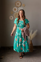 Woman in a teal floral dress standing in a room with decorative wall art and a plant.