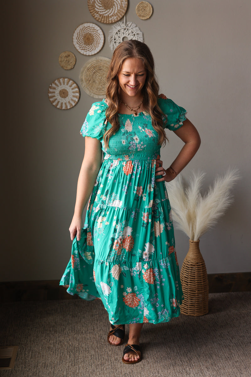 Woman in a teal floral dress standing in a room with decorative wall art and a vase.