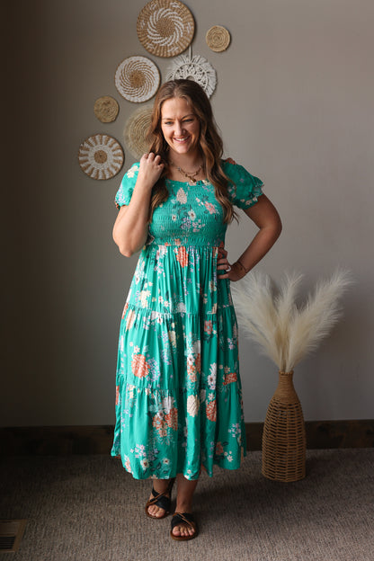 Woman wearing a teal floral dress standing in a room with decorative wall art and a vase.