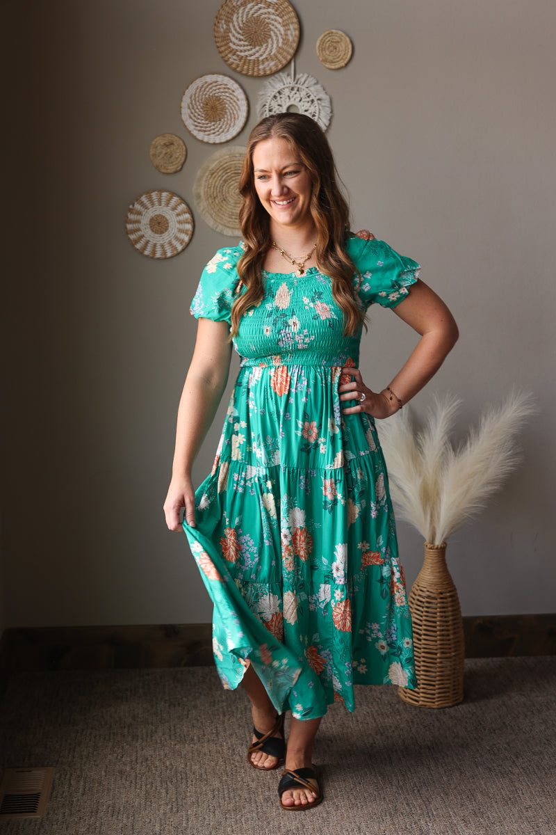 Woman in a teal floral dress standing in a room with decorative wall art and a vase.