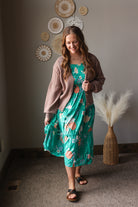 Woman in a floral dress and brown cardigan standing in a room with decorative wall art and a vase.