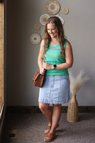 Woman in a green top and denim skirt standing indoors with decorative elements on the wall.