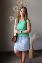 Woman in a green tank top and denim skirt standing indoors with decorative elements on the wall.