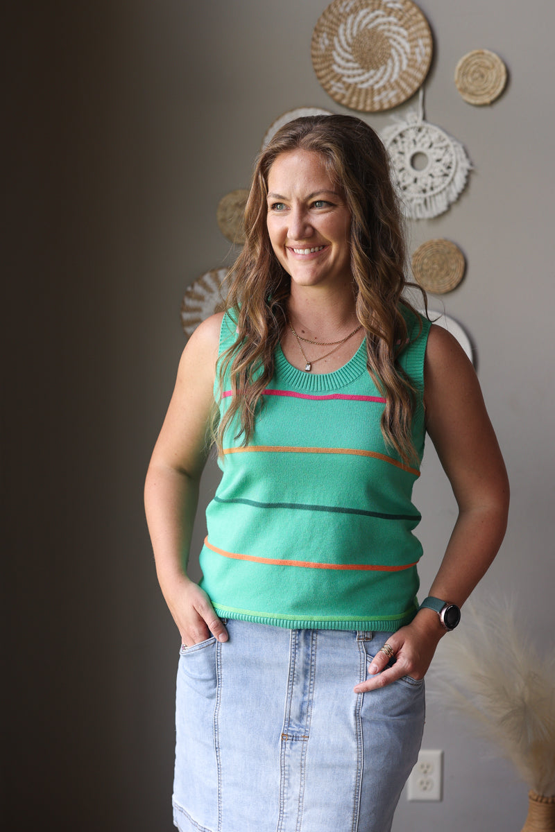 Woman wearing a green striped tank top and blue jean skirt standing against a neutral wall with decorative items.